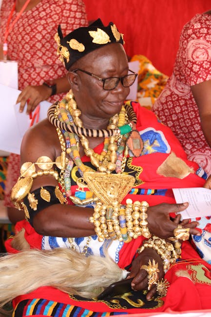The image features an older woman dressed in traditional, vibrant attire with elaborate gold jewelry, including necklaces, bracelets, and a headpiece, set against a red backdrop. She is seated, holdin