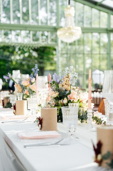 A floral arrangement featuring a variety of fresh flowers including lavender, soft pink roses, white and blush peonies, and purple irises, displayed atop a white table setting in a sunlit conservatory