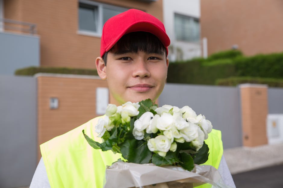 A young delivery person wearing a red cap and neon yellow vest holds a bouquet of white roses with green leaves, wrapped in white tissue paper, in front of a modern residential building with brick and