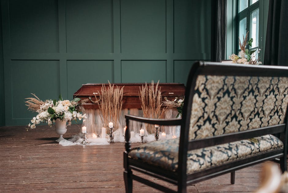 A floral arrangement consisting of white roses, baby's breath, and green foliage placed in a white ceramic vase on a dark wooden table. The arrangement is complemented by dried pampas grass behind it,